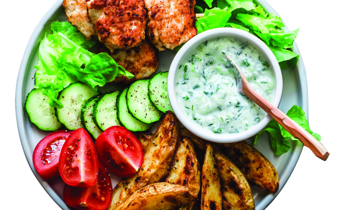 Fish skewers, baked potatoes, vegetables and yogurt greens sauce on dark background, top view